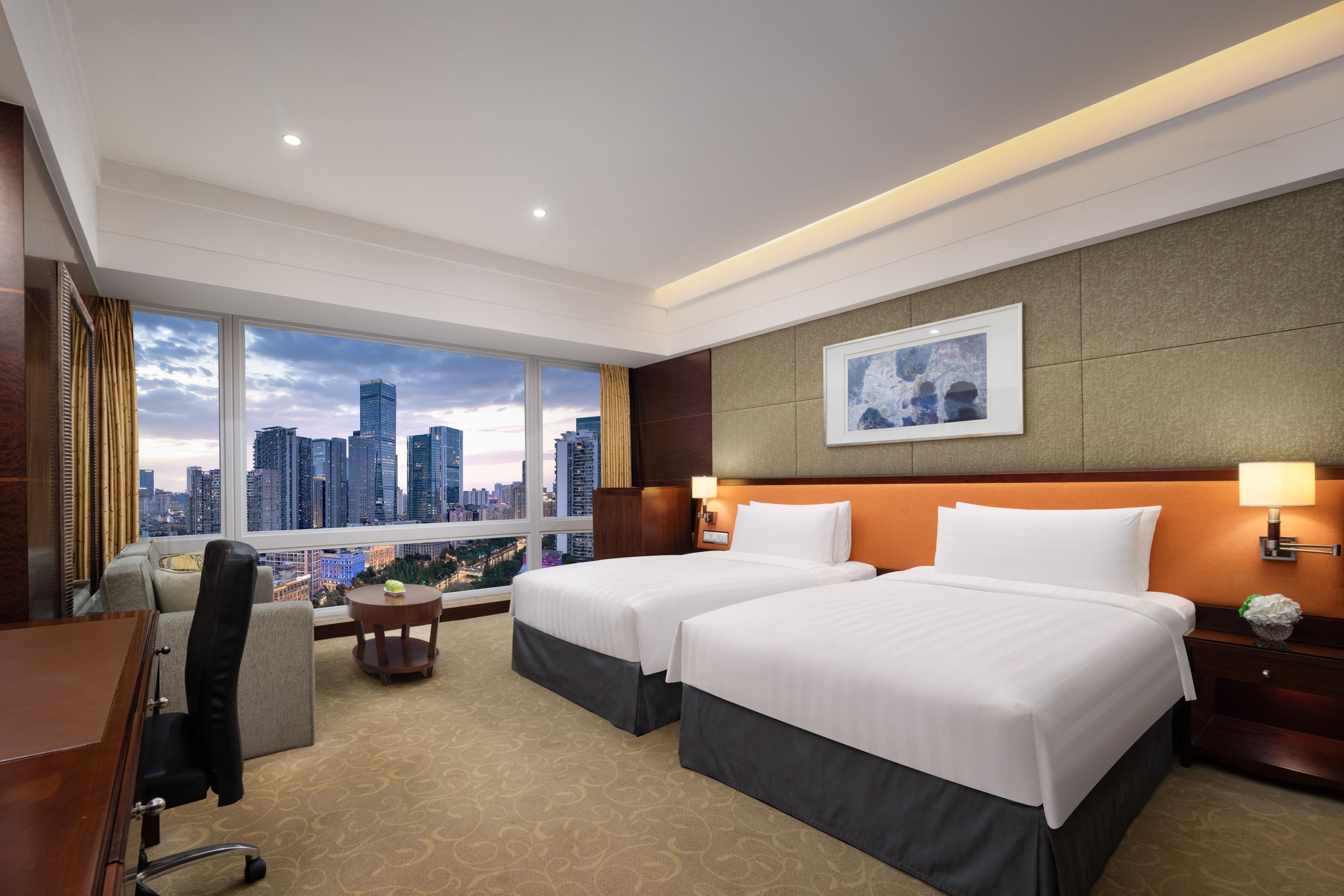 A hotel room with two beds, a desk, a chair, a view of the cityscape, and a painting on the wall.