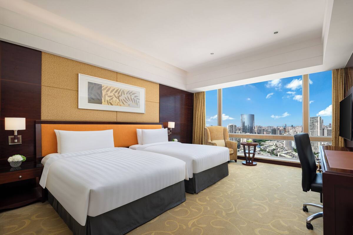 Habitación de hotel con dos camas, un escritorio, una silla, un televisor y un ventanal con vista a la ciudad.