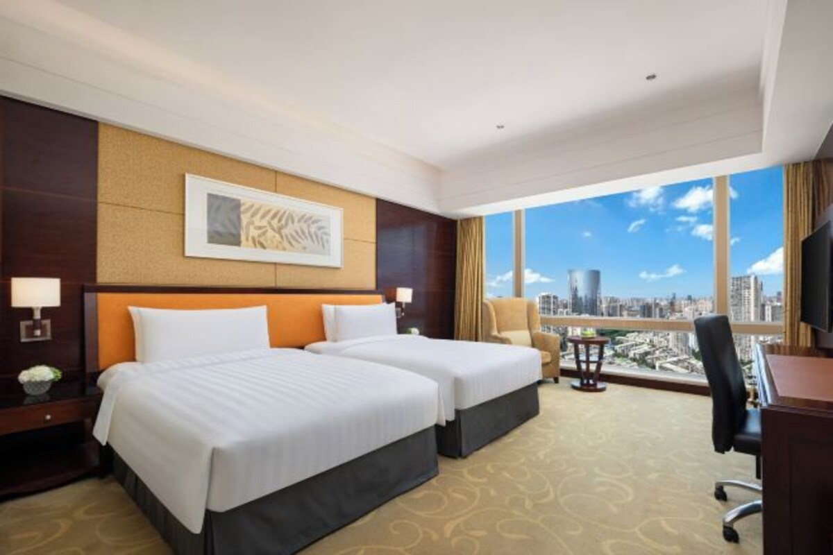 Habitación de hotel con dos camas, un escritorio, una silla y un amplio ventanal con vistas a la ciudad.