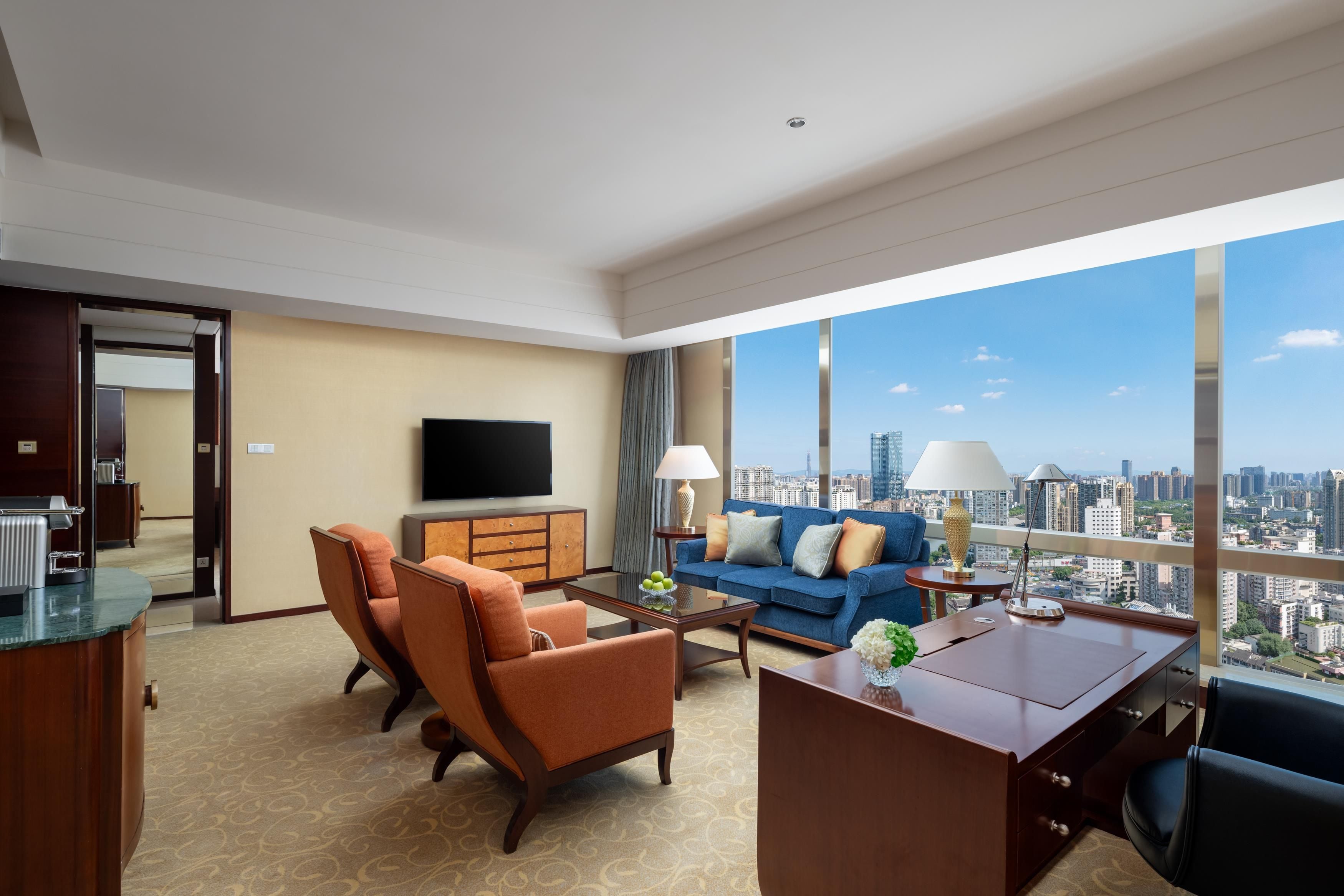 Une chambre d’hôtel moderne avec vue sur la ville, un téléviseur à écran plat, un canapé, deux fauteuils, une table basse et un bureau.