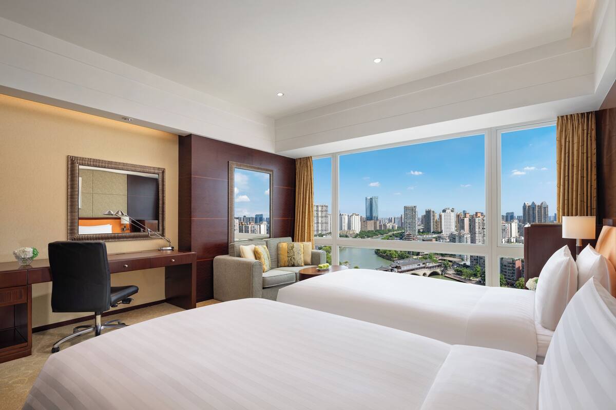 Habitación de hotel con dos camas, un escritorio, una silla y un amplio ventanal con vista a la ciudad.