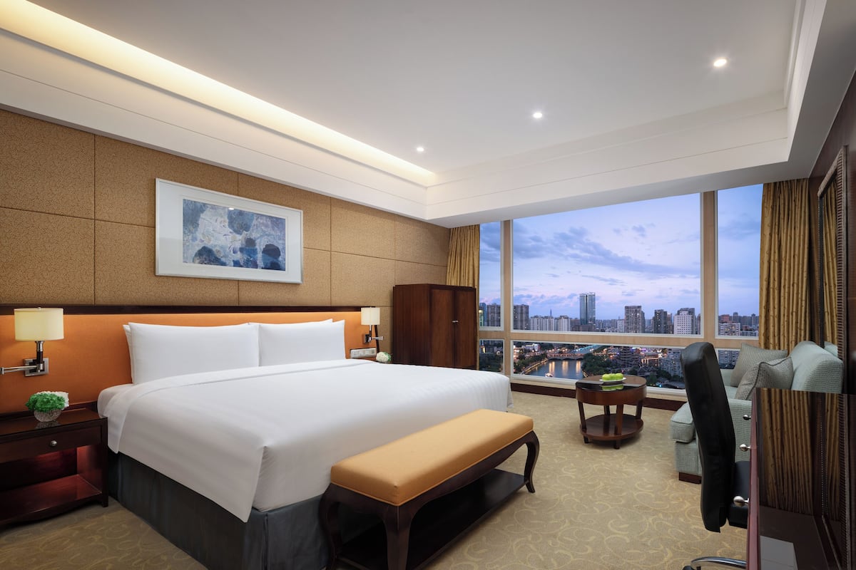 Habitación de hotel con una cama grande, un banco, un escritorio con lámpara, vista panorámica de la ciudad a través de amplias ventanas y un cuadro en la pared.