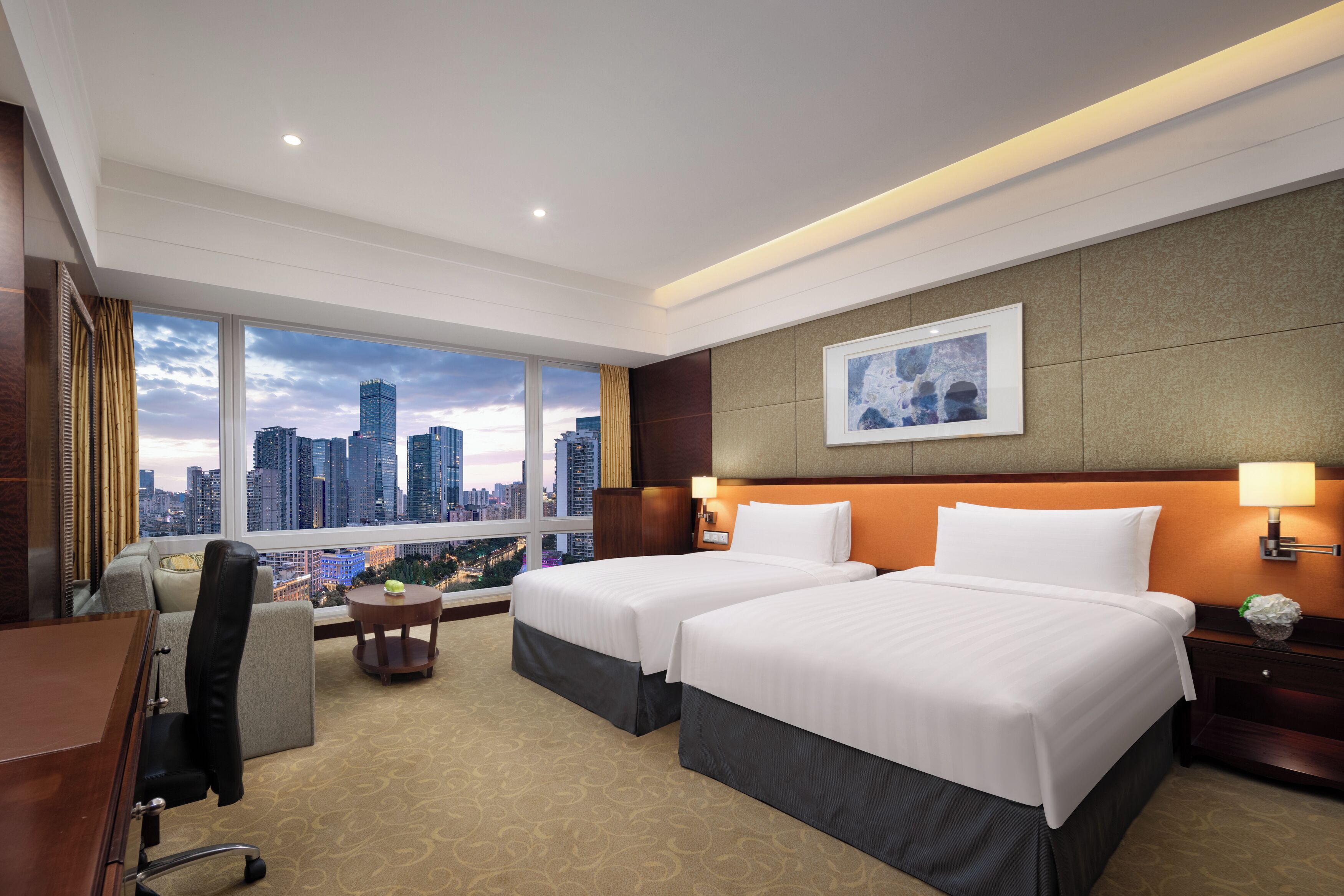A hotel room with two beds, a desk, a chair, a small table with a lamp, and a view of the cityscape.