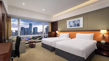 A hotel room with two beds, a desk, a chair, a small table with a lamp, and a view of the cityscape.