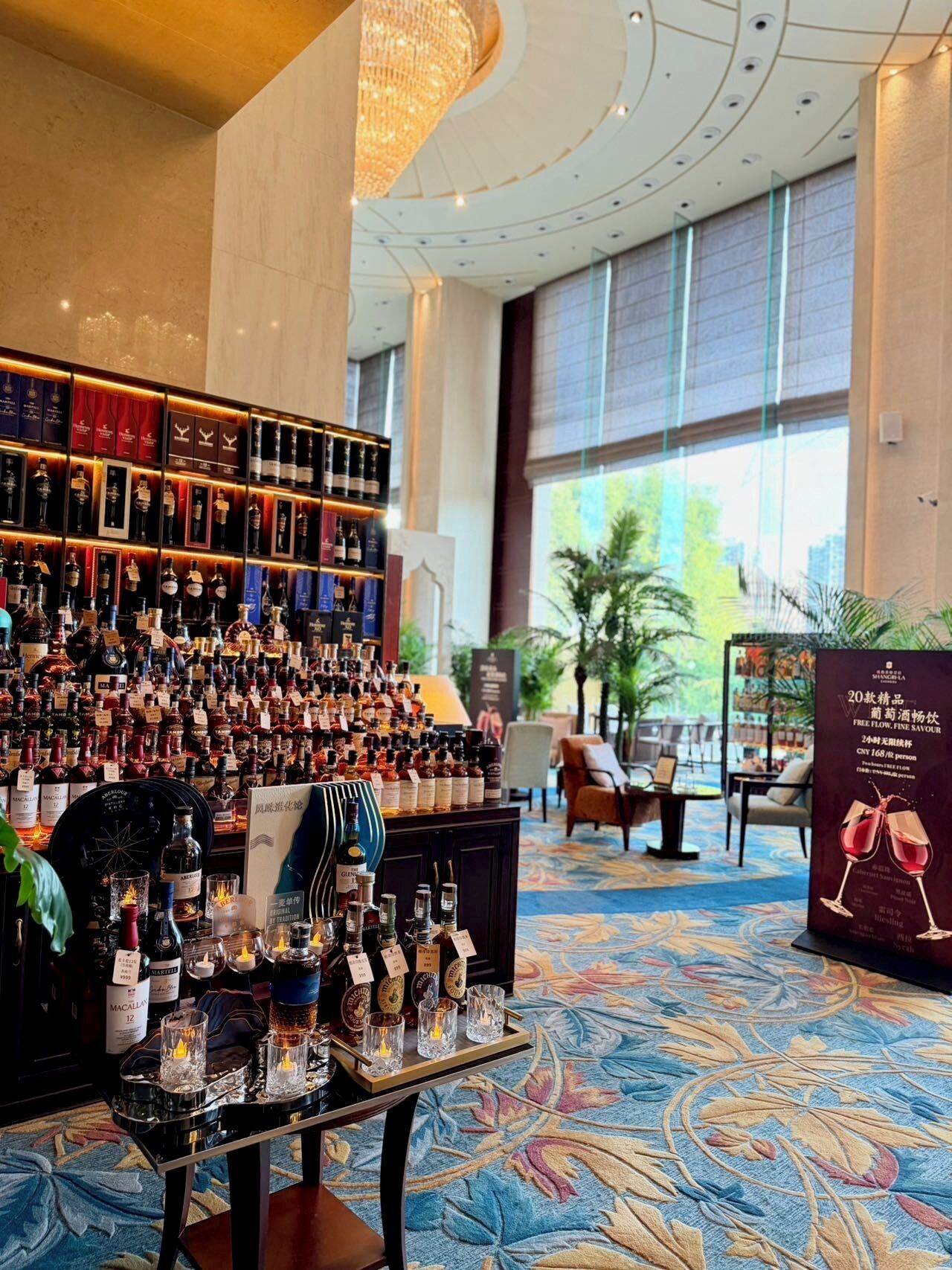 A bar area with a variety of liquor bottles and glasses, a chandelier, and a large window with a view of greenery.