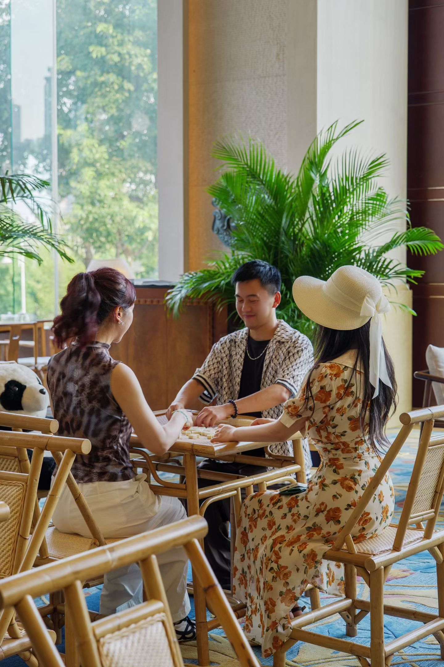 A group of people seated at a table in a room with large windows, wooden chairs, and potted plants.