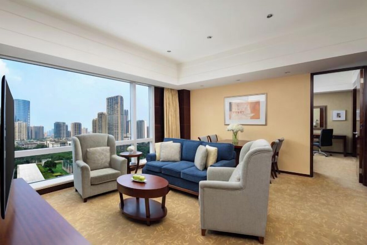 A modern hotel room with a city view, a sofa, armchairs, a coffee table, and a TV.