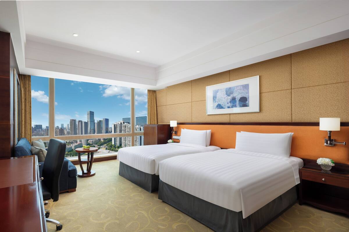 Habitación de hotel con dos camas, un escritorio, una silla, una mesita y un ventanal con vistas a la ciudad.