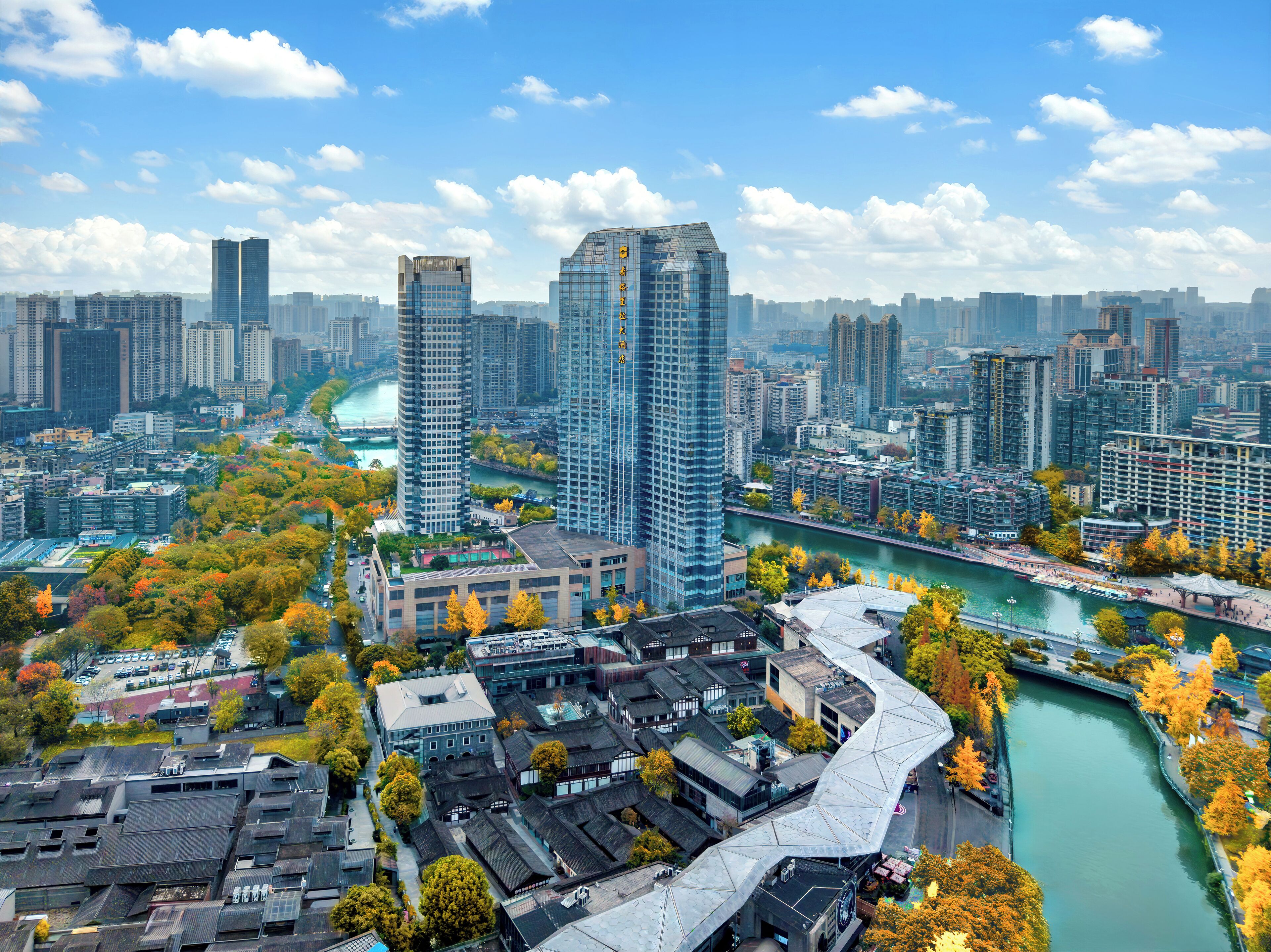 A high-rise building with a modern glass facade, surrounded by a mix of traditional and contemporary architecture, near a river with a pedestrian walkway.