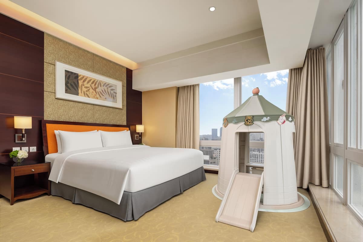 Habitación de hotel con una cama grande, una mesita de noche con lámpara, una obra de arte en la pared, una ventana con cortinas y una casita de juegos con tobogán.