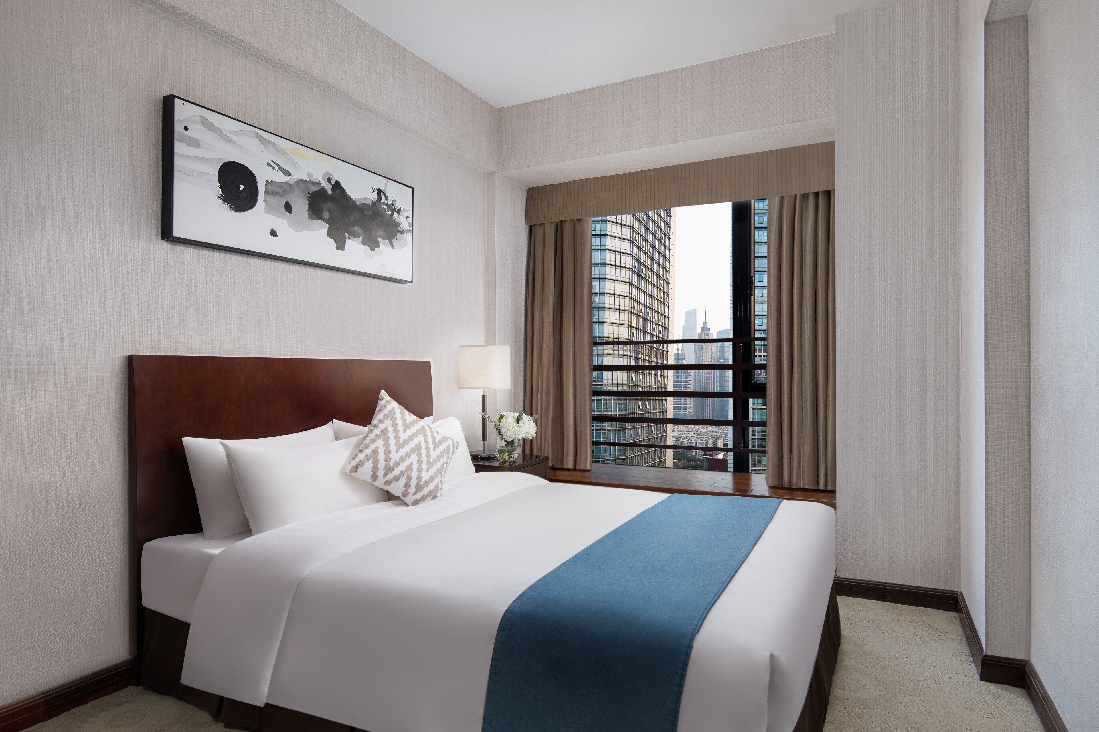 A hotel room with a large bed, a city view, a framed artwork on the wall, and a nightstand with a lamp.