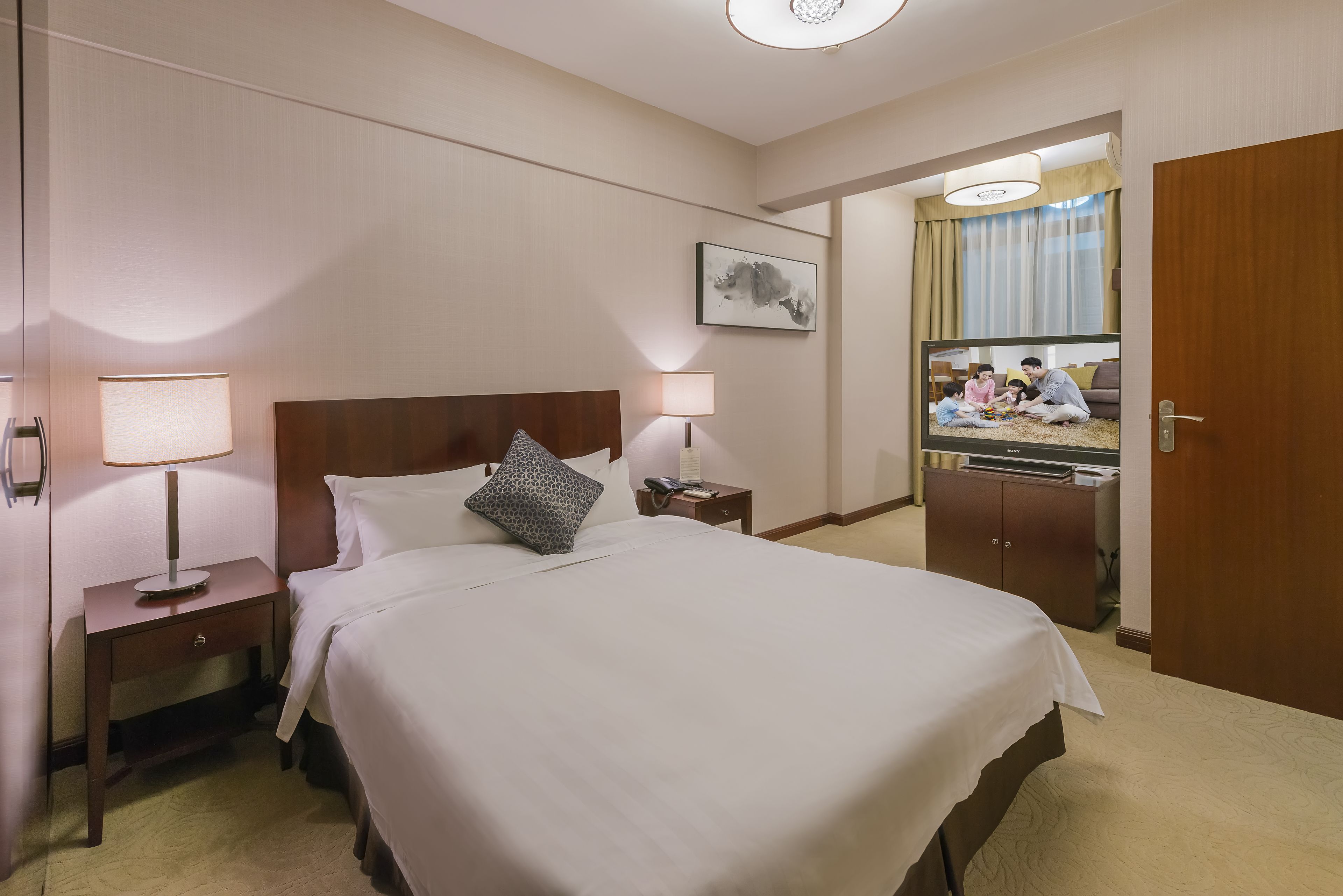 Une chambre d’hôtel avec un grand lit, deux tables de chevet avec des lampes, une tête de lit en bois, un tableau au mur et une vue sur une famille dans la chambre voisine.