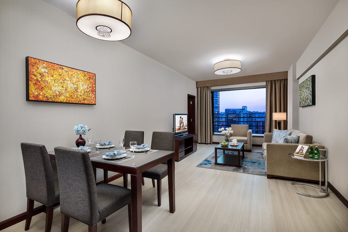 A modern hotel room with a dining area, a living area with a sofa and armchair, a TV, and a large window with a city view.