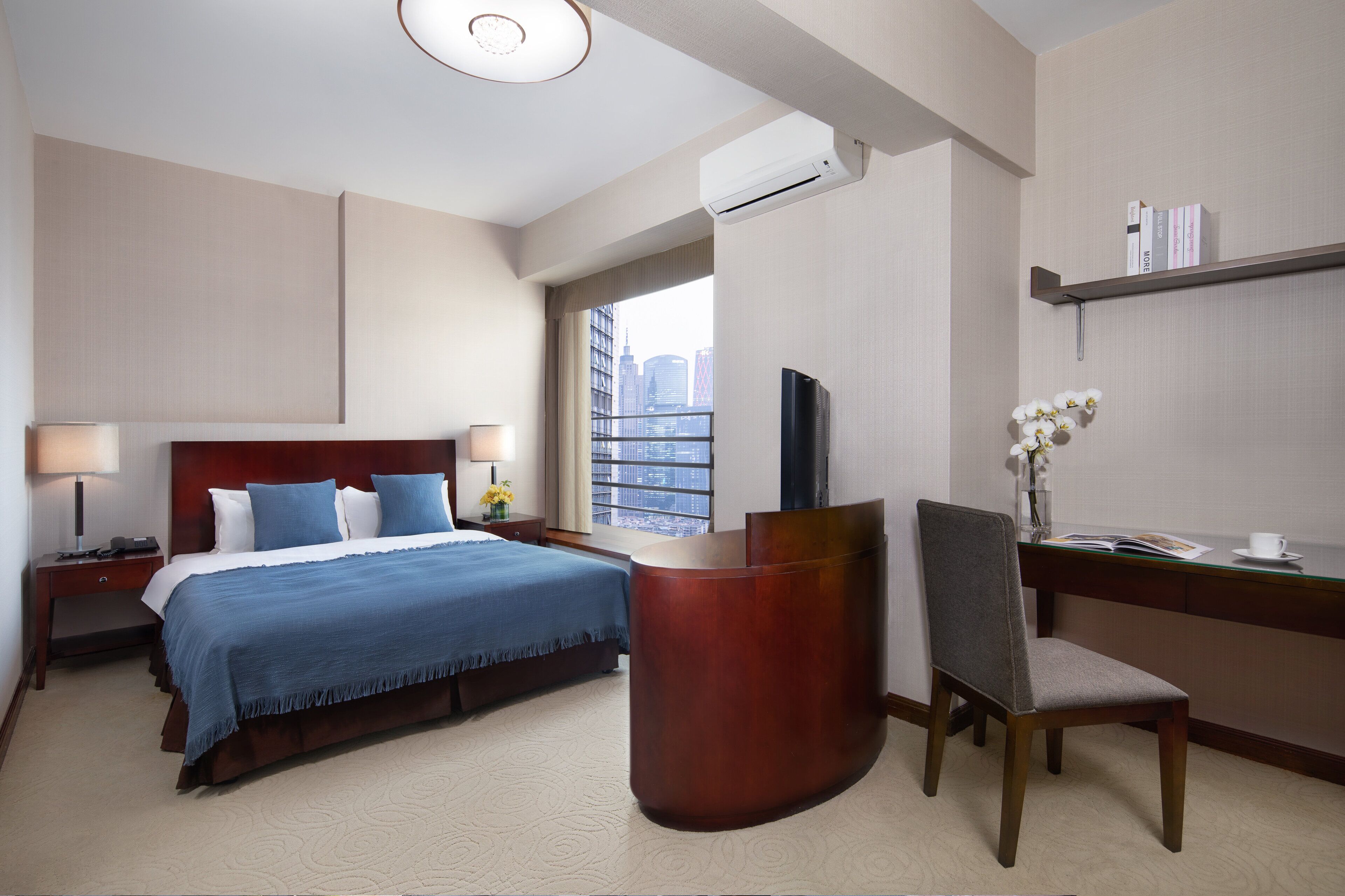 A hotel room with a bed, bedside tables, a desk, a chair, a TV, and a window with a city view.