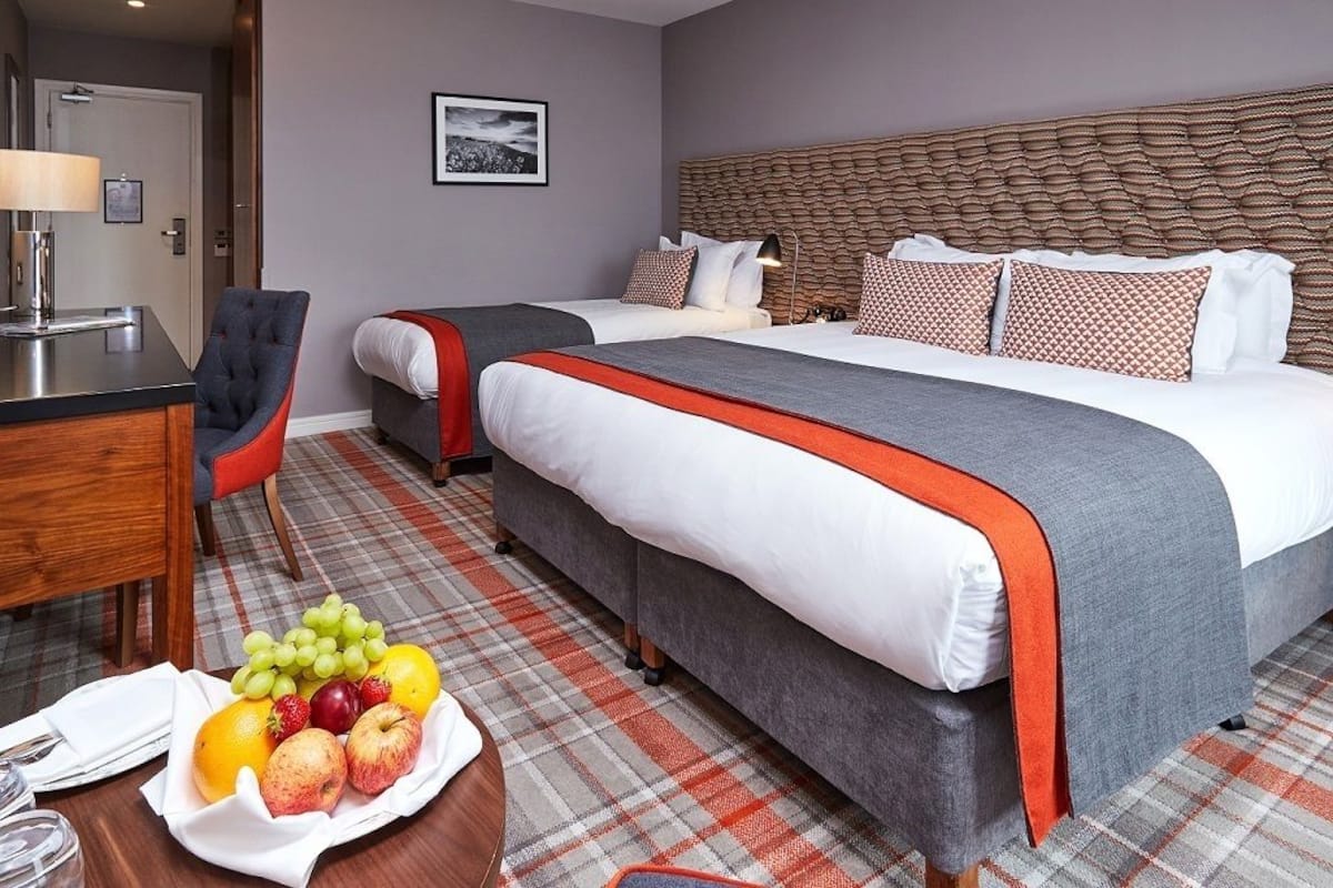 Ein Hotelzimmer mit einem großen Bett, einem kleineren Bett, einem Schreibtisch mit Stuhl und einem Obstkorb auf dem Boden.