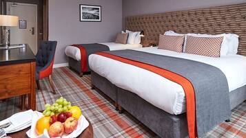 A hotel room with a large bed, a smaller bed, a desk with a chair, and a fruit basket on the floor.