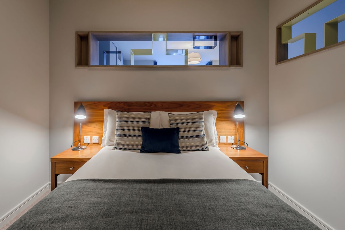 Ein ordentlich eingerichtetes Hotelzimmer mit einem großen Bett, zwei Nachttischlampen, einem Kopfteil mit Spiegel und Blick ins Zimmer durch ein Fenster.