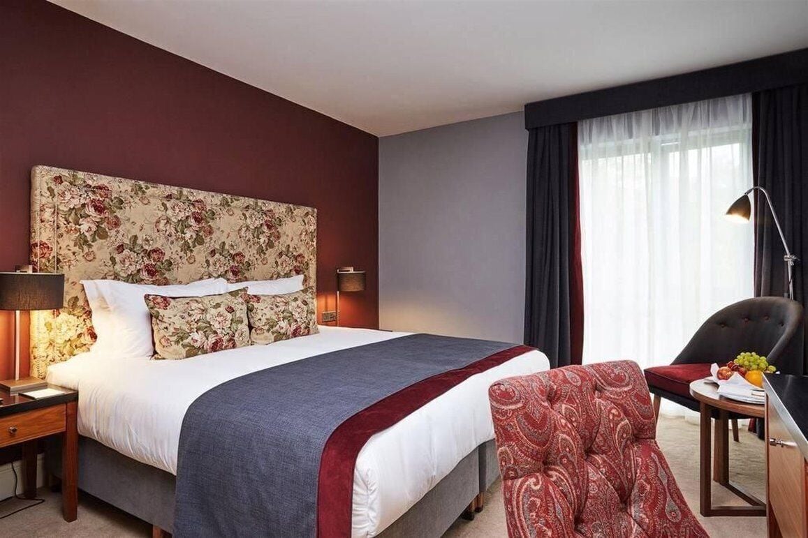 Una camera d'albergo con un letto grande, una scrivania con una lampada, una sedia e una finestra con tende.