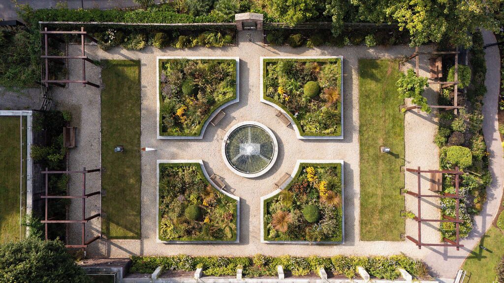 An aerial view of a landscaped garden with a central circular fountain, surrounded by four rectangular flower beds, and a gravel path.