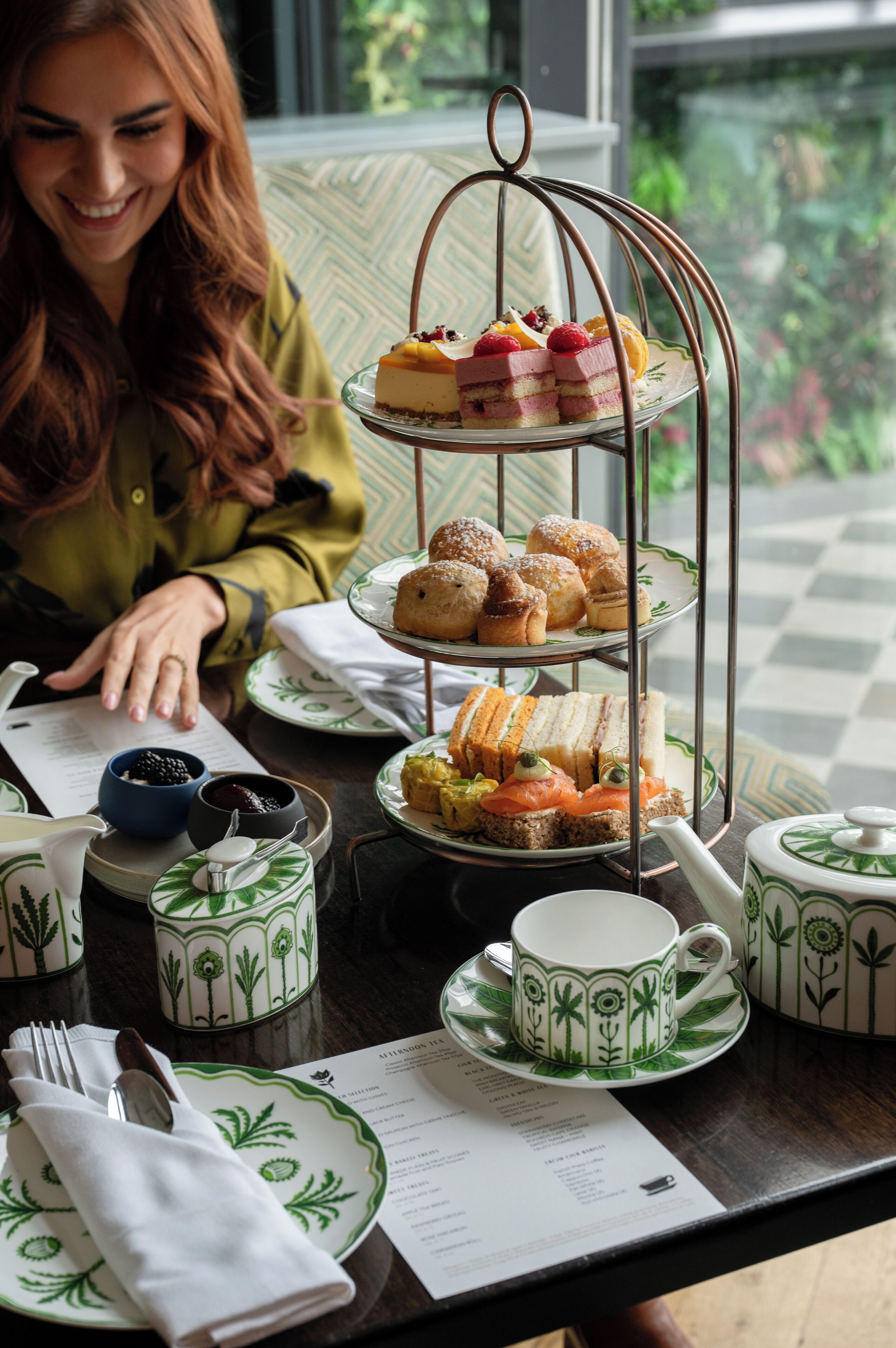 A tiered dessert stand with various pastries and sandwiches, a tea set, and a menu on a table with a view of greenery outside.