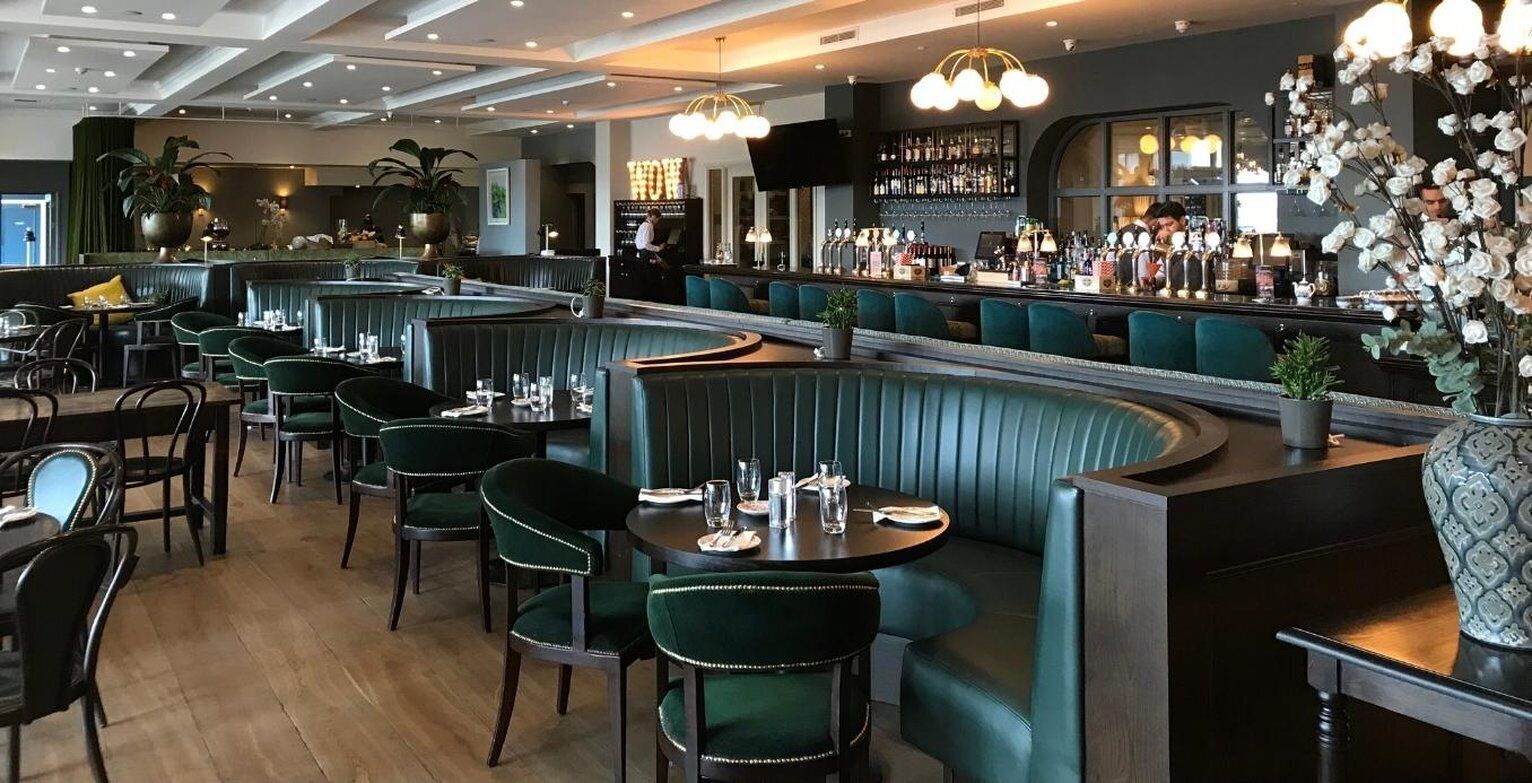 Un restaurante con cabinas y mesas, una zona de bar con un bartender, y una variedad de plantas y decoración.