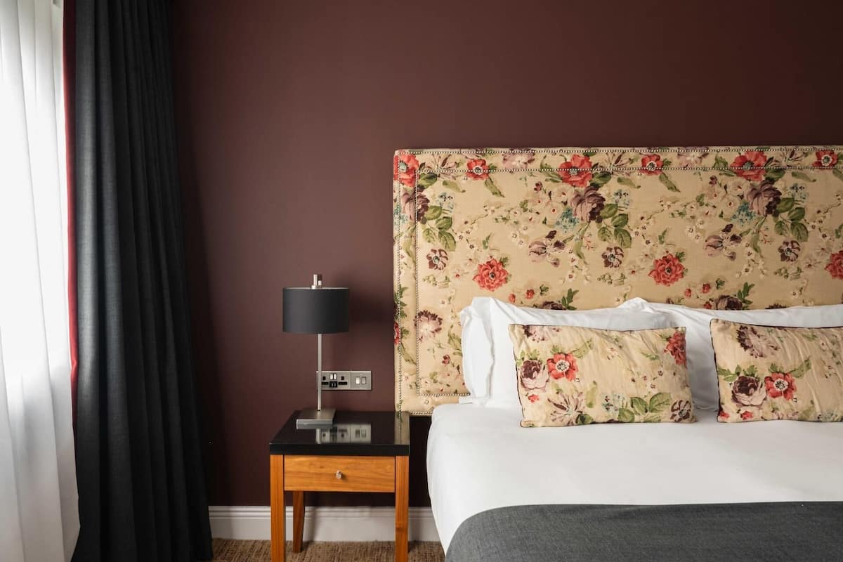 Ein Hotelzimmer mit einem blumengemusterten Kopfteil, einem Nachttisch mit Lampe und einem Bett mit weißer Bettwäsche.