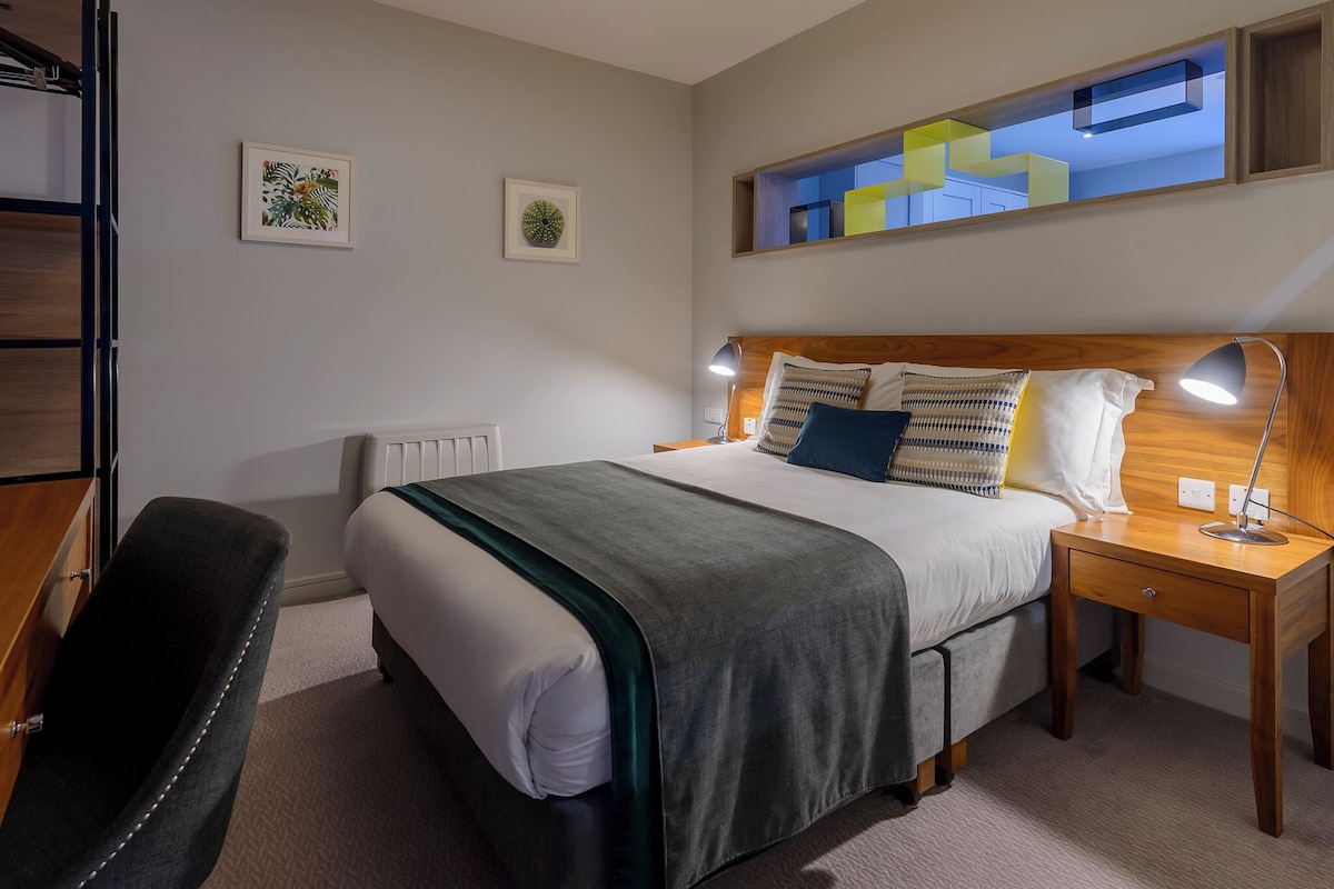 Un dormitorio ordenado con cama, mesita de noche, lámpara y una silla.