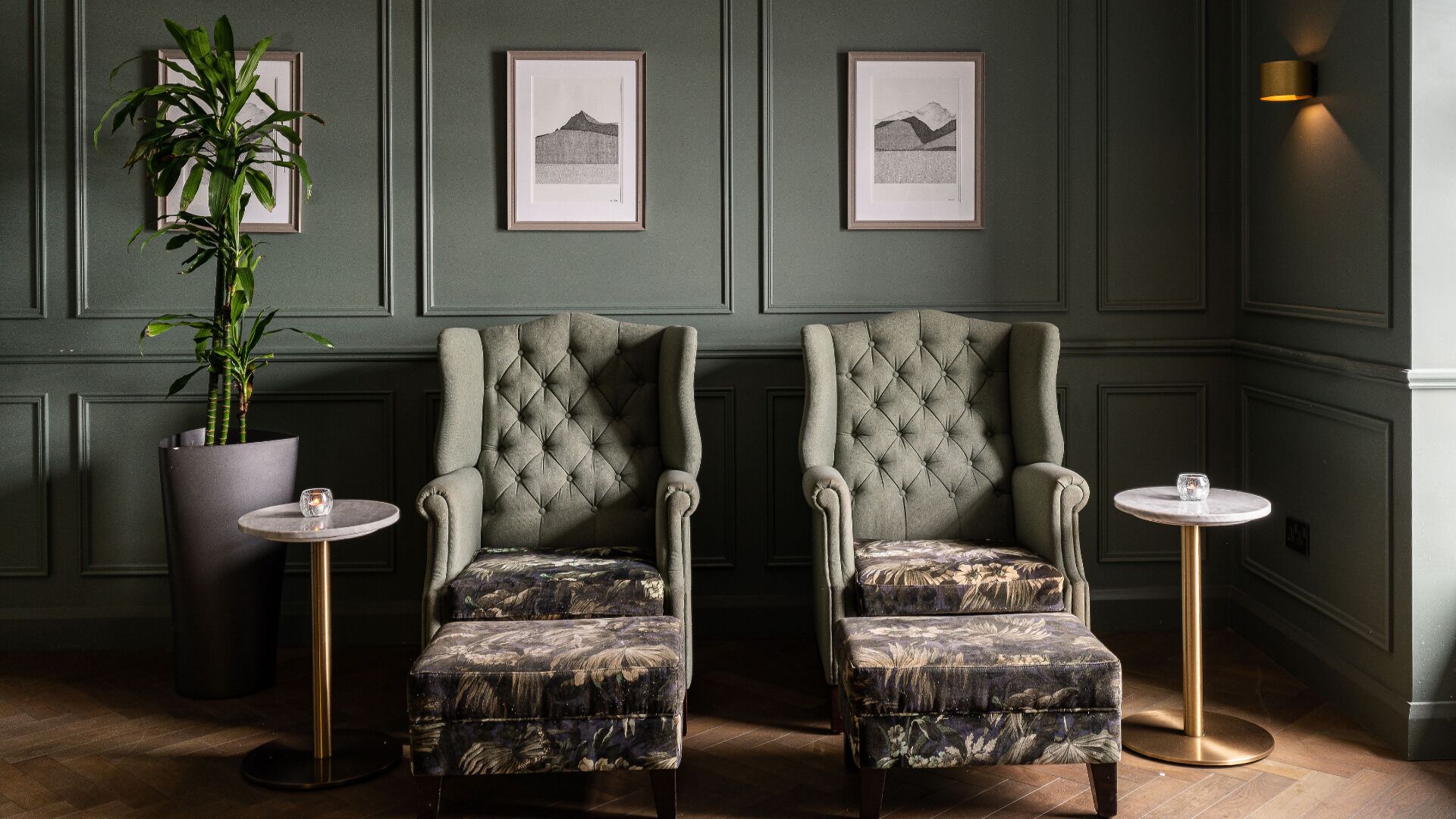 A room with two upholstered armchairs, a potted plant, framed artwork on the wall, and a small table with a glass.