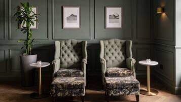 A room with two upholstered armchairs, a potted plant, framed artwork on the wall, and a small table with a glass.