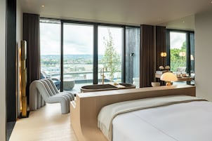 Una habitación de hotel moderna con una cama grande, zona de estar y vistas a un paisaje urbano.