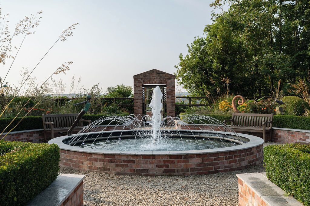 A circular brick fountain with water jets, surrounded by a brick wall and benches, set in a landscaped garden with trees and shrubs.