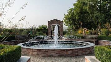 A circular brick fountain with water jets, surrounded by a brick wall and benches, set in a landscaped garden with trees and shrubs.