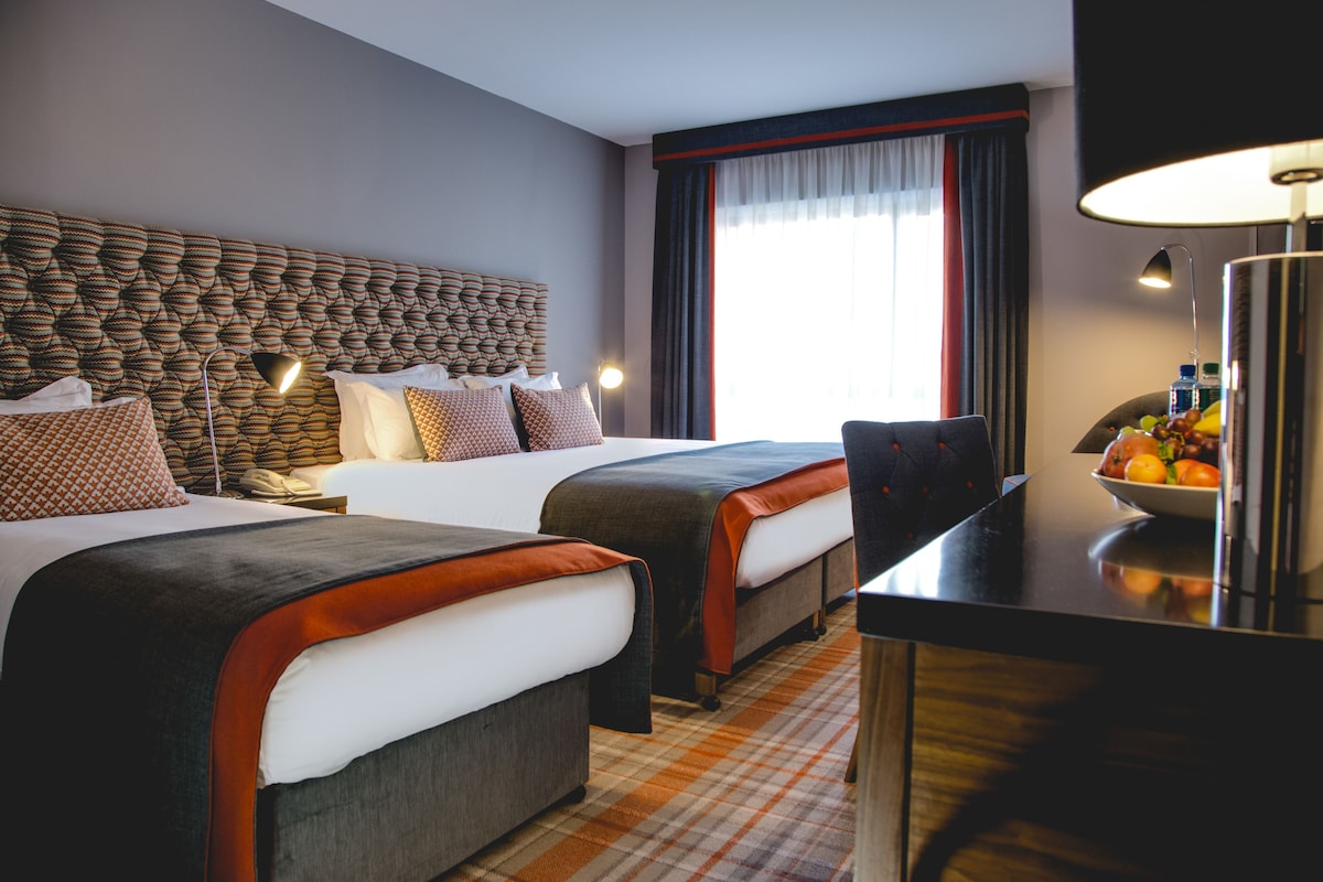 Habitación de hotel con dos camas, cabeceros, lámparas de noche, un escritorio y un frutero.