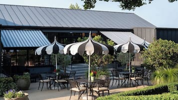 Un patio al aire libre con mesas y sillas, rodeado de plantas en macetas y un edificio con techo de metal.