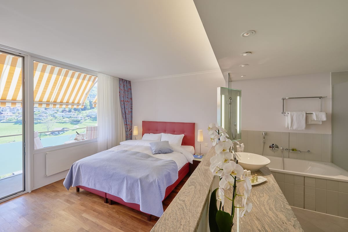 A modern hotel room with a large bed, a bathroom with a bathtub, and a view of a green landscape.