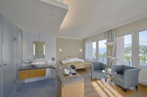 Ein modernes Hotelzimmer mit einem großen Bett, einer Sitzecke mit Sofa und kleinem Tisch, einem Badezimmer mit Waschbecken und Spiegel sowie großen Fenstern mit Blick auf die Berge.