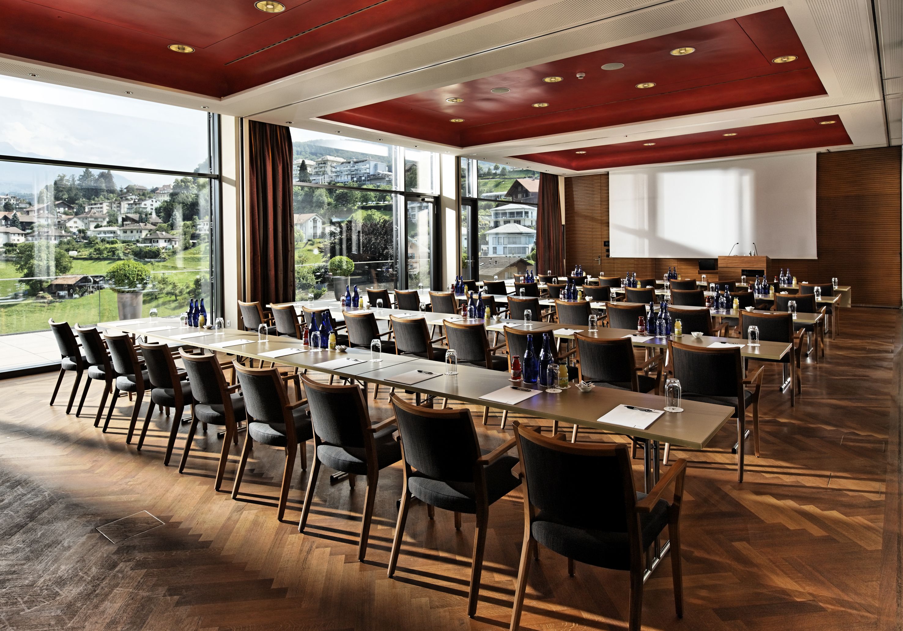 A conference room with multiple tables and chairs, large windows with a view of a town, and a wooden floor.