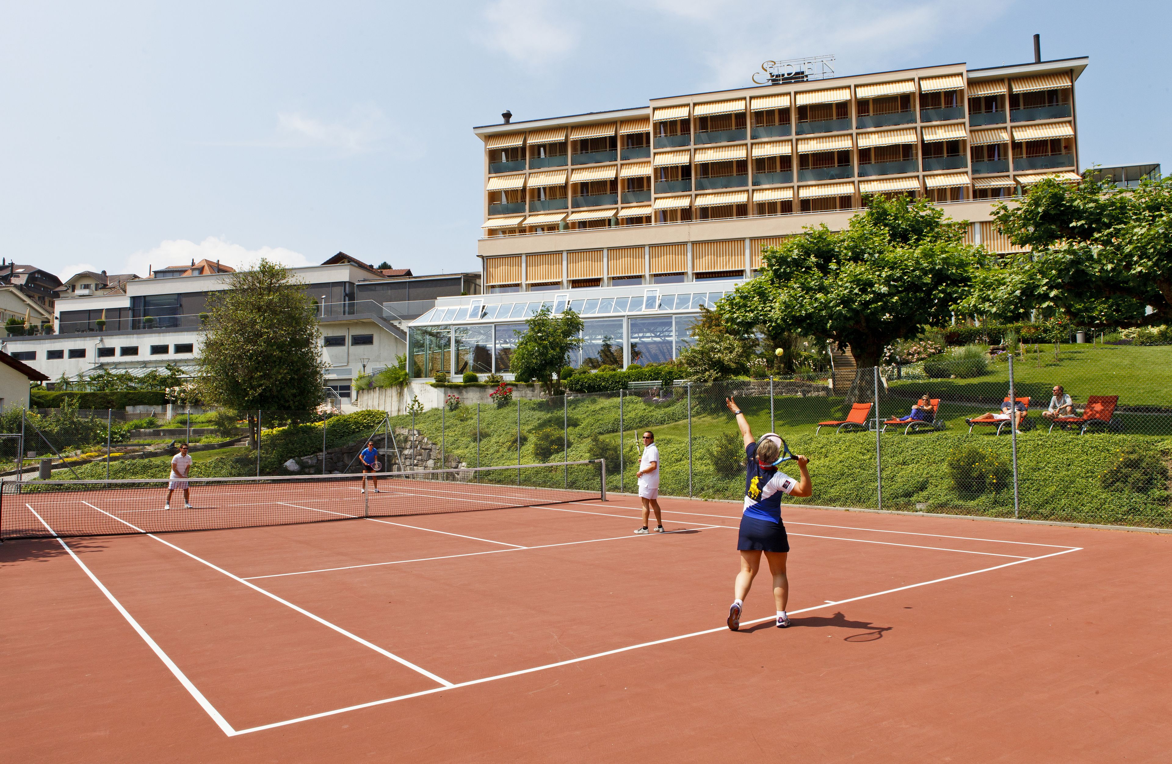 A tennis court with players, surrounded by buildings and greenery.