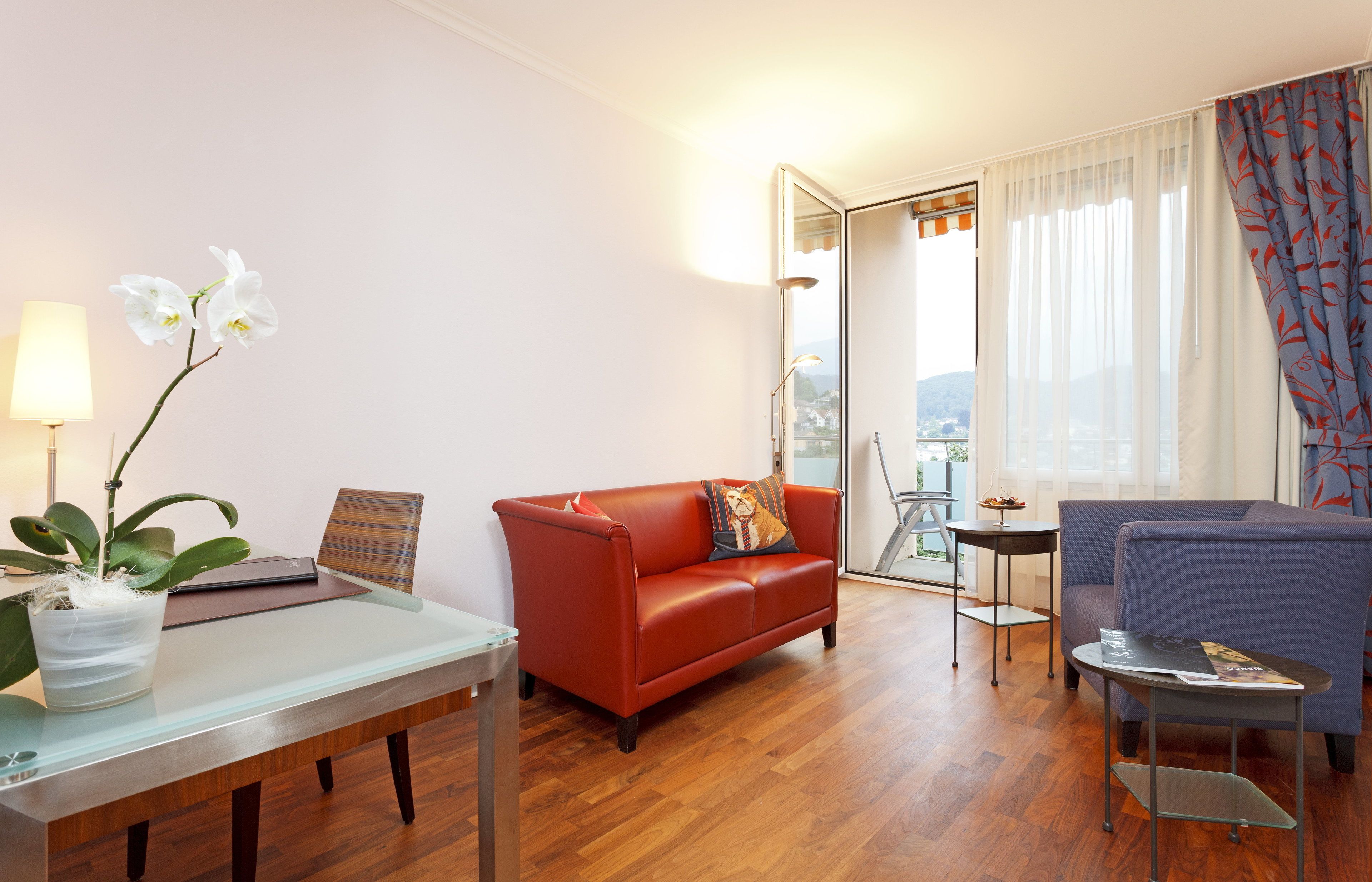 Un salón moderno con una mesa de vidrio, sofá rojo, sillón azul y vistas de la ciudad a través de amplias ventanas.