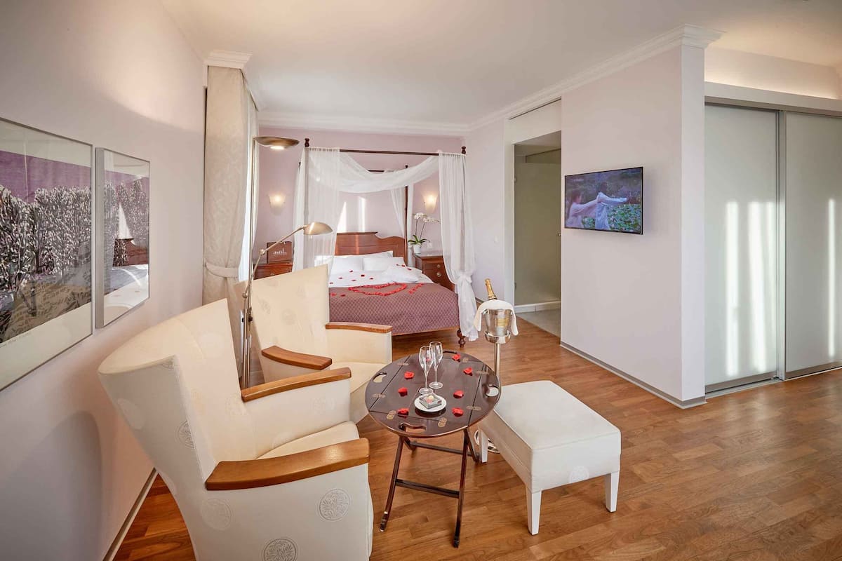 A bedroom with a canopy bed, a sitting area with a table set for two, and a large window.