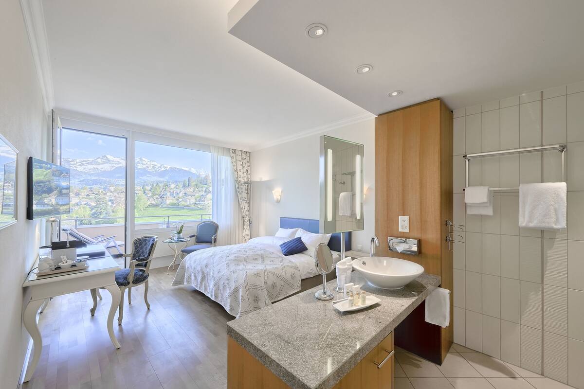 A modern hotel room with a large bed, a desk with a laptop, a bathroom with a large sink and mirror, and a view of the mountains through the window.