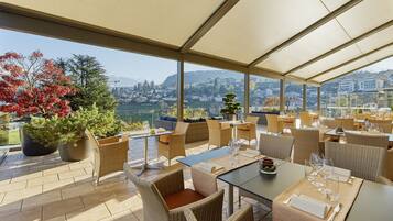 Un restaurante con terraza, grandes ventanales y vistas a la ciudad y a las montañas.