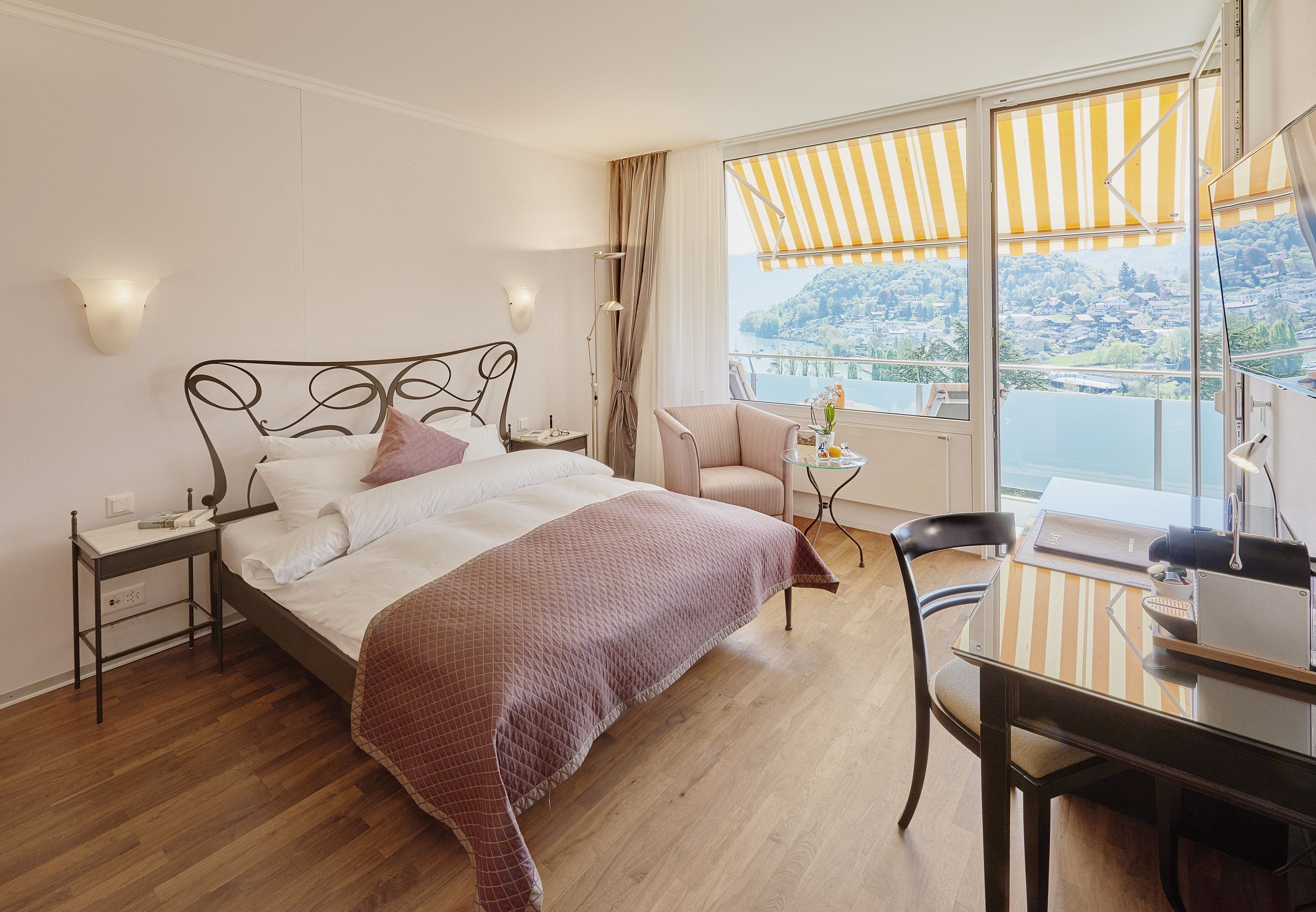 A hotel room with a bed, a desk, a chair, a small table, and a view of a lake and hills.