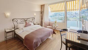A hotel room with a bed, a desk, a chair, a small table, and a view of a lake and hills.