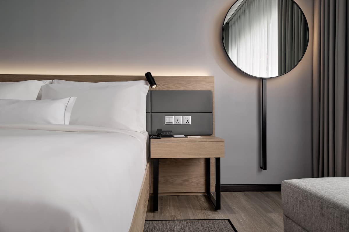 A modern hotel room with a neatly made bed, a bedside table with a lamp, a round mirror, and a power outlet.