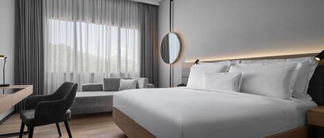 A modern hotel room with a large bed, a desk with a chair, and a view of the city through large windows.