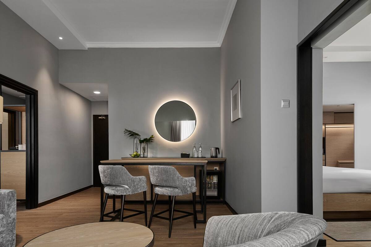 A modern hotel room with a wooden desk, two grey upholstered chairs, a round mirror, and a bed with a wooden headboard.