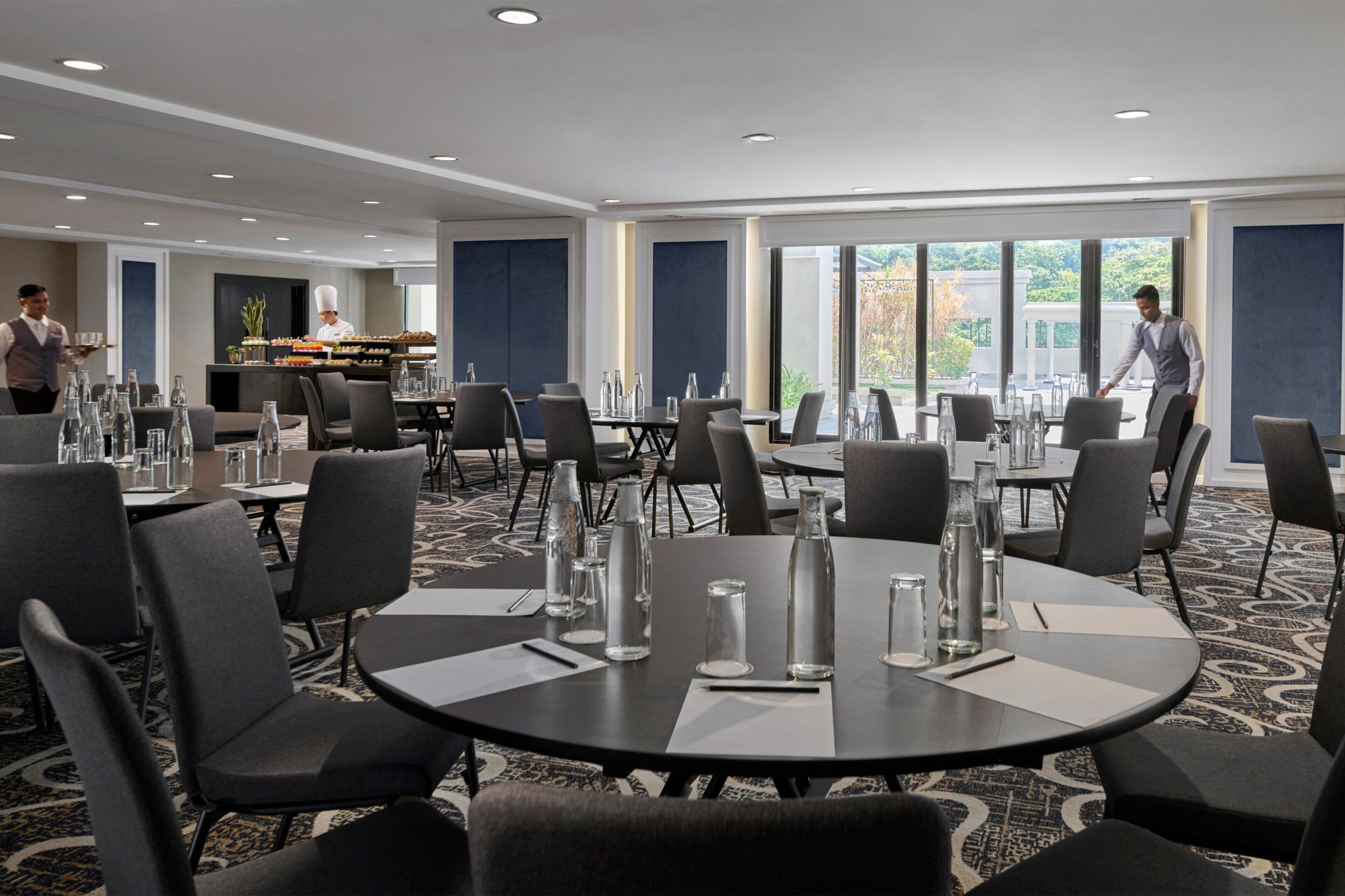 A modern conference room with round tables, grey chairs, and a buffet area in the background.