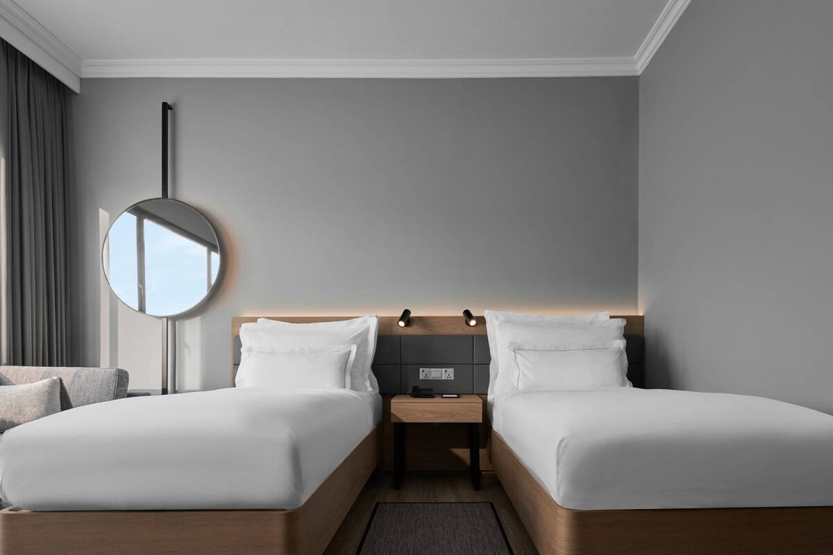 A modern hotel room with two single beds, a bedside table with a lamp, a round mirror, and a view of the sky through the window.