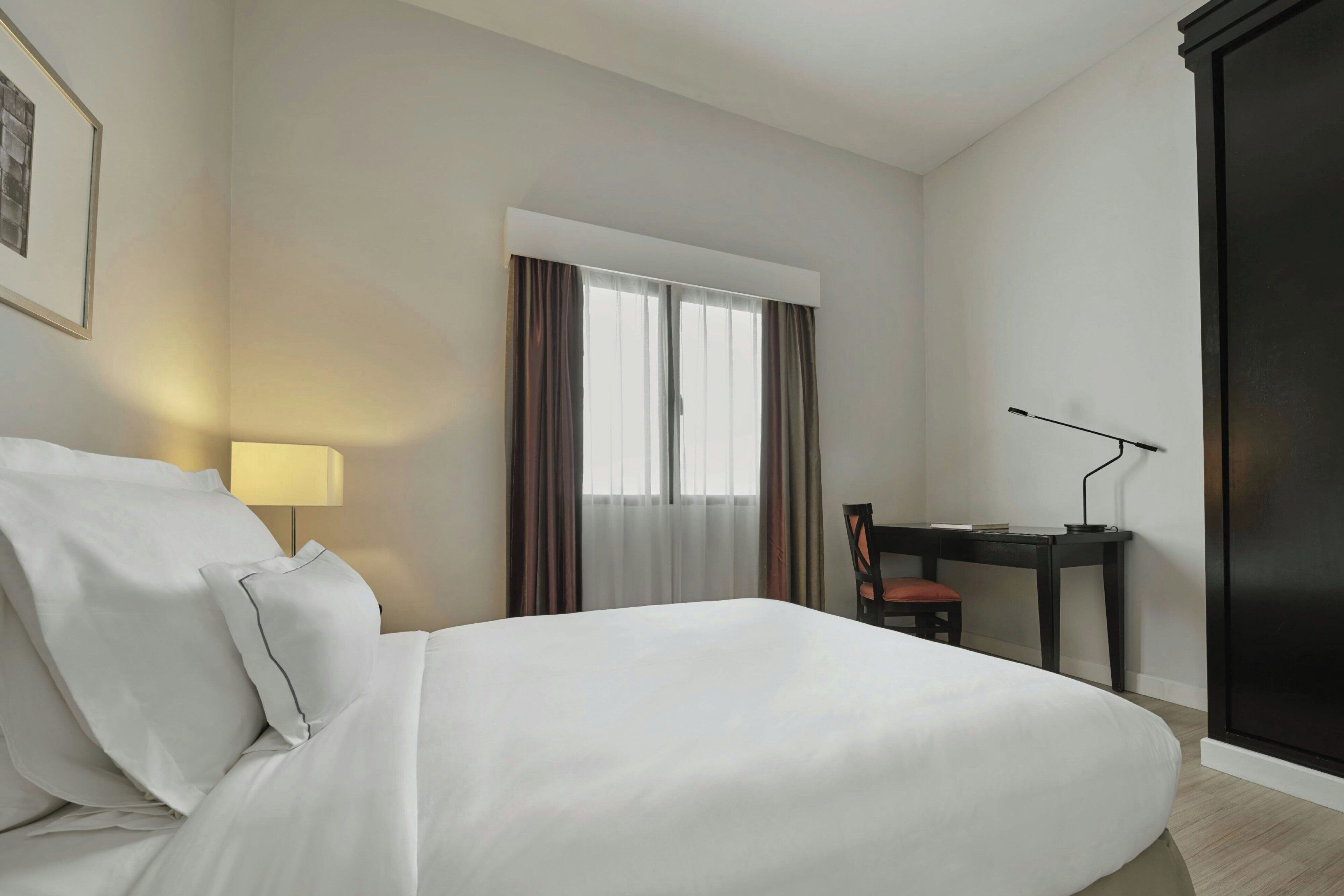 A hotel room with a bed, a desk, a chair, a lamp, and a window with curtains.